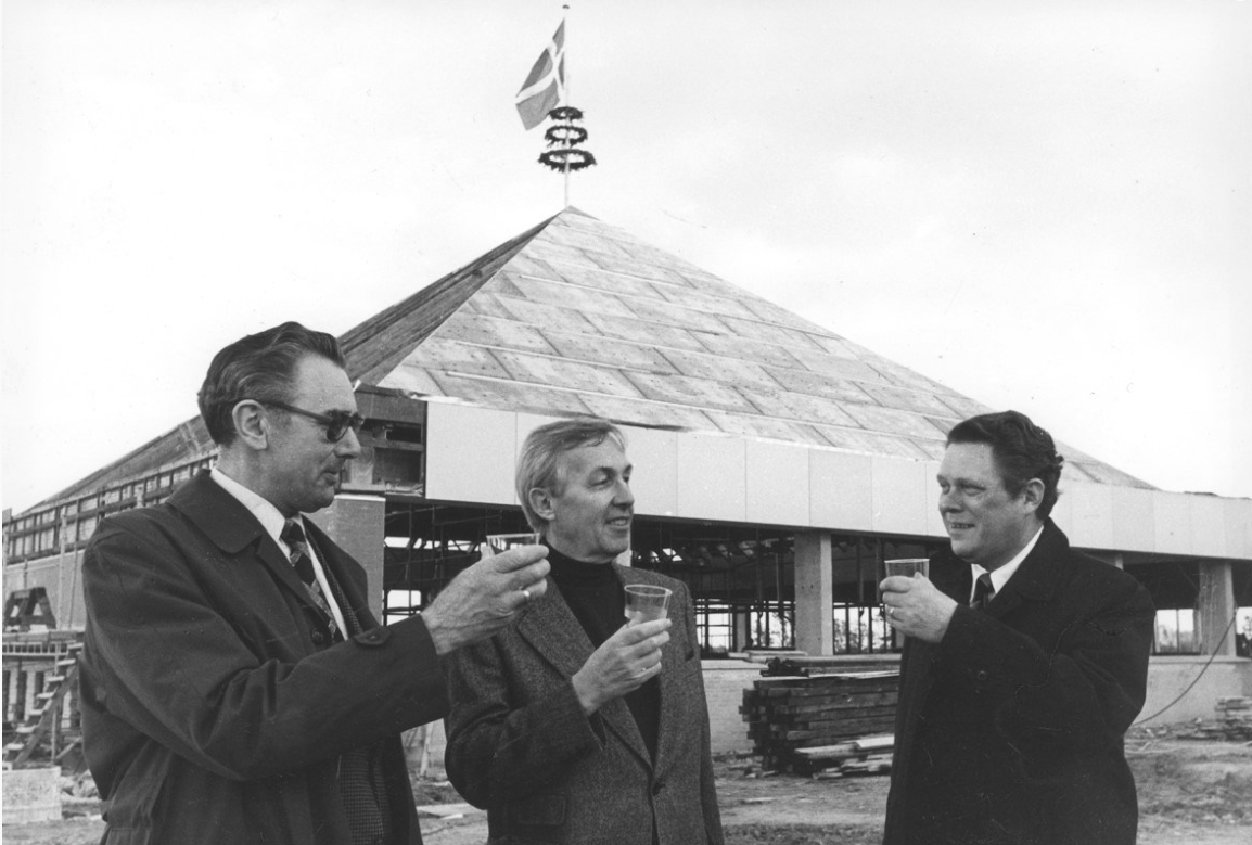 Rejsegildet i oktober 1977 af den første version af Hollænderhallens Svømmehal med den genkendelige pyramide over ”den dybe ende” af idrætsbassinet. Fra venstre daværende borgmester Albert Svendsen, arkitekt Paul Grønning og svømmeklubbens mangeårige formand Tage Welander. (Arkiv.dk)
